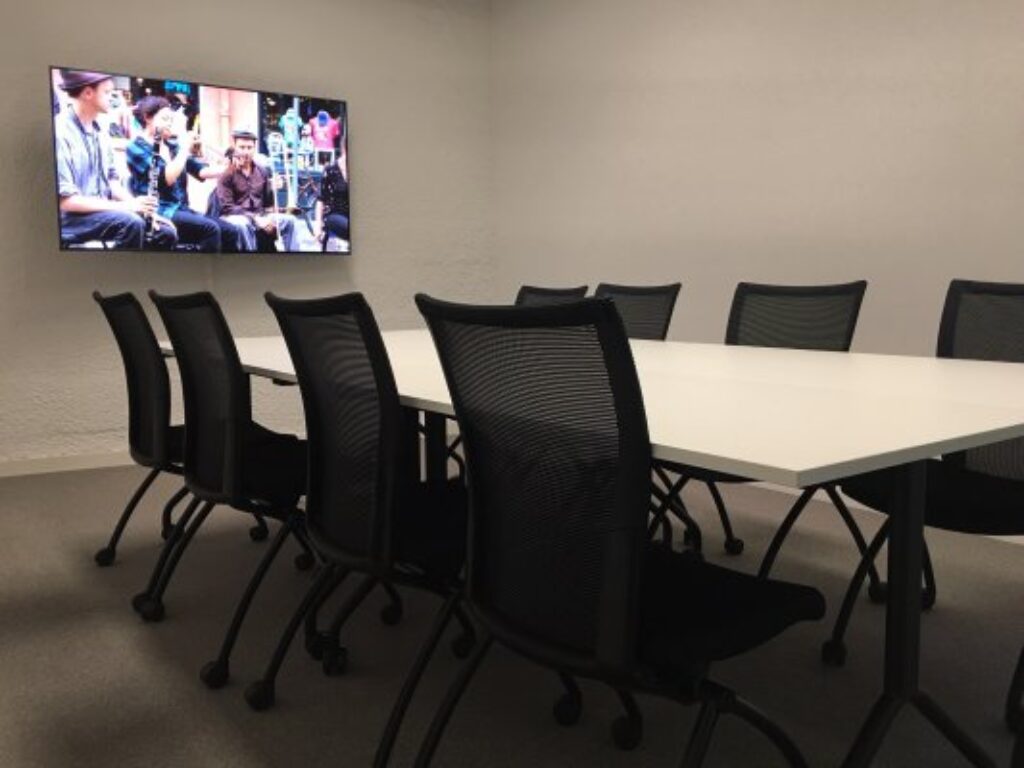 Une table entouré de huit chaises, avec contre le mur en écran. Permet de faire des conférences